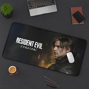 Może przedstawiać: Czarna prostokątna podkładka pod mysz z białym napisem "RESIDENT EVIL requiem" i wizerunkiem mężczyzny. Na podkładce znajduje się biała mysz komputerowa. Widoczny jest również laptop, ołówki i filiżanka kawy.