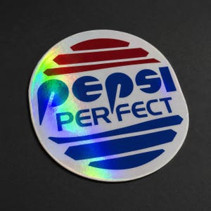May include: A circular holographic sticker featuring the text PEPSI PERFECT in bold blue letters. The design includes a red horizontal striped graphic above the text and blue horizontal striped graphics below, creating a retro-futuristic aesthetic.