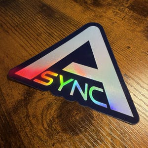 May include: A triangular sticker featuring a dark blue background with a large, stylized letter A in a holographic, reflective finish. The word SYNC is printed in a matching holographic font across the center of the triangle, creating a vibrant, multi-colored light effect against the dark border.