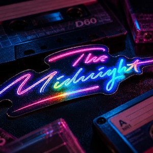 May include: A die-cut sticker featuring the text The Midnight in a vibrant neon-style gradient of pink, purple, blue, and yellow. The sticker is surrounded by several black plastic audio cassette tapes with visible labels and reels in a dark setting.