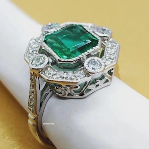 Peut inclure: Une bague en argent ornée d'une grande pierre verte de taille émeraude au centre, entourée de plus petites pierres précieuses transparentes. La bague présente un design orné d'inspiration Art déco avec des détails complexes et une finition polie.