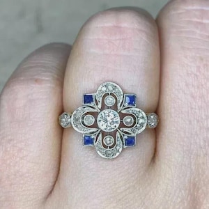 May include: A silver-toned ring featuring a central round diamond surrounded by a decorative metal frame with four smaller round diamonds, four square-cut blue sapphires, and intricate metalwork detailing in a symmetrical floral-inspired pattern.