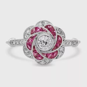Könnte beinhalten: Ein silberfarbener Ring mit einem zentralen runden weißen Edelstein, umgeben von einem blumenförmigen Heiligenschein aus geschwungenen, tiefroten Rubinen und kleinen, klaren runden Diamanten. Die Ringschiene ist mit einer Reihe winziger Diamanten besetzt.