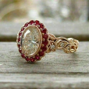 Könnte beinhalten: Ovaler Diamantring mit rotem Edelstein-Halo. Der Ring hat ein goldenes Band mit aufwendigen Blatt- und Diamantakzenten. Der Ring liegt auf einer Holzoberfläche.