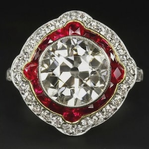 Puede incluir: Un anillo con una gran piedra central redonda y facetada, rodeada por un halo festoneado de rubíes rojos vibrantes. Todo el engaste está enmarcado por un borde de pequeños diamantes blancos brillantes montados en una banda de metal plateado.