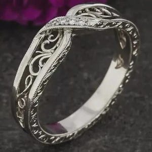 May include: A silver-toned ring featuring an intricate twisted band design with openwork scroll patterns and a row of small, clear, round-cut stones embedded along the top section of the band, showcasing a polished metallic finish throughout.