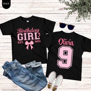 Op de afbeelding: Twee zwarte t-shirts met roze tekst. Eén zegt "Birthday GIRL EST. 2017" met een strik. De andere zegt "Olivia 9". Een blauwe spijkerbroek, witte sneakers en een zonnebril staan ook op de foto.