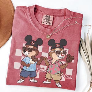 Peut inclure: T-shirt rouge Comfort Colors avec un motif de dessin anim&eacute; de Tic et Tac portant des lunettes de soleil et tenant des glaces. Le t-shirt a un fond &agrave; carreaux et les mots "Chip" et "Dale" sont imprim&eacute;s.