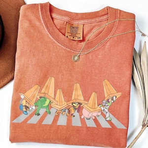 May include: A coral-coloured t-shirt featuring a graphic of cartoon characters crossing a zebra crossing, each wearing a traffic cone. The shirt has a crew neck and short sleeves. A gold necklace with a pendant is visible.