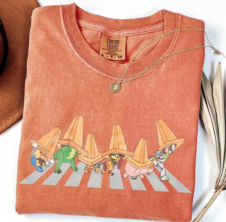 May include: A coral-coloured t-shirt featuring a graphic of cartoon characters crossing a zebra crossing, each wearing a traffic cone. The shirt has a crew neck and short sleeves. A gold necklace with a pendant is visible.