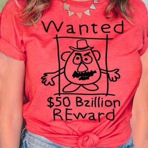 K&ouml;nnte beinhalten: Ein korallenfarbenes T-Shirt mit einer schwarz-wei&szlig;en Cartoon-Illustration eines kartoffelartigen Charakters in einem Fahndungsplakat. Der Text "Wanted" und "$50 Bzillion REWARD" sind ebenfalls in Schwarz dargestellt.