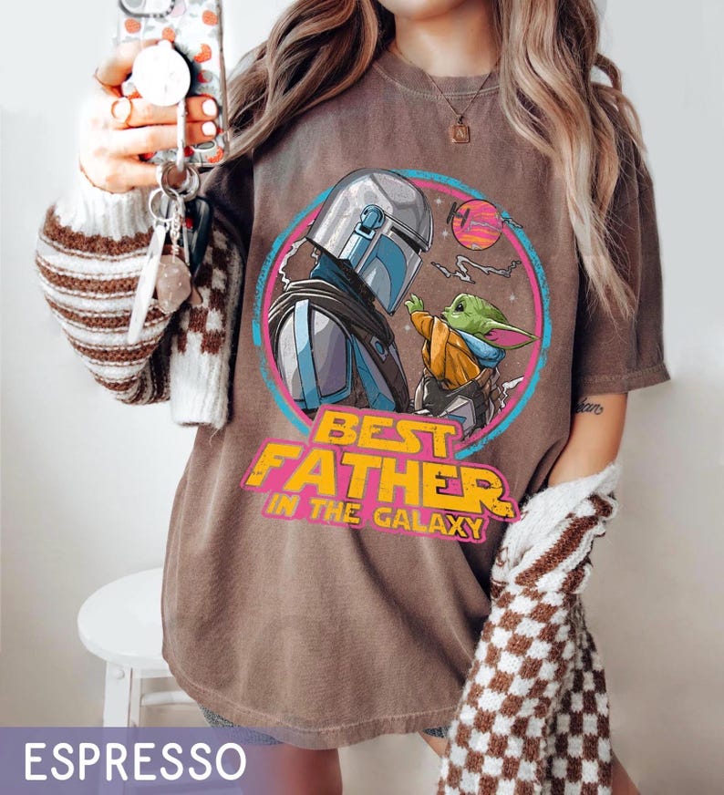 May include: Espresso-colored t-shirt featuring a graphic of The Mandalorian and Grogu, with the text "Best Father in the Galaxy" in yellow and pink. The design is set within a circular frame.