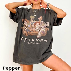 May include: Pepper-colored t-shirt featuring a graphic of characters from the Toy Story movie, seated on a couch. The word "FRIENDS" is printed in white, with "SINCE 1995" below. The shirt has short sleeves and a relaxed fit.