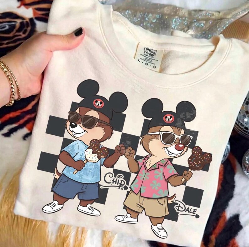 Peut inclure: Sweat-shirt cr&egrave;me avec une illustration de dessin anim&eacute; de Tic et Tac portant des lunettes de soleil et des chapeaux Mickey, tenant des glaces. Le motif comprend un motif &agrave; carreaux noir et blanc et les mots "Chip" et "Dale".