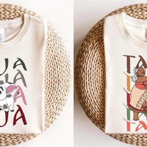 May include: Two cream-colored t-shirts with graphic designs. One features a cartoon pig with the word "PUA" in gray, pink, and maroon. The other shirt displays a woman dancing with the word "TALA" in brown, yellow, green, and maroon.