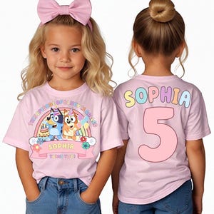 Puede incluir: Dos camisetas rosas. Una presenta un dibujo animado con el texto "This episode of Bluey is called" y "Sophia turns two". La otra tiene el nombre "Sophia" y el número "5" en la espalda. El nombre está en colores pastel.