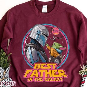 May include: Maroon sweatshirt featuring a graphic of The Mandalorian and Grogu, with the text "BEST FATHER IN THE GALAXY." The design is set within a pink and blue circular frame, with a sunset background.