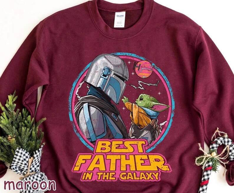 May include: Maroon sweatshirt featuring a graphic of The Mandalorian and Grogu, with the text "BEST FATHER IN THE GALAXY." The design is set within a pink and blue circular frame, with a sunset background.