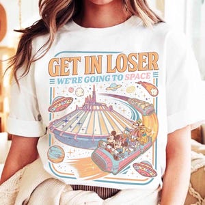 May include: White t-shirt with a colorful graphic featuring a space-themed design. The graphic includes the text "GET IN LOSER WE'RE GOING TO SPACE" above a roller coaster with Disney characters, planets, and UFOs. The design is in shades of orange, blue, and pink.