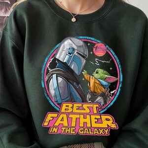 May include: Dark green sweatshirt featuring a colorful graphic of a character in silver armor and a small green creature. The graphic is framed by a pink and blue circle with the text "BEST FATHER IN THE GALAXY" in yellow and orange.