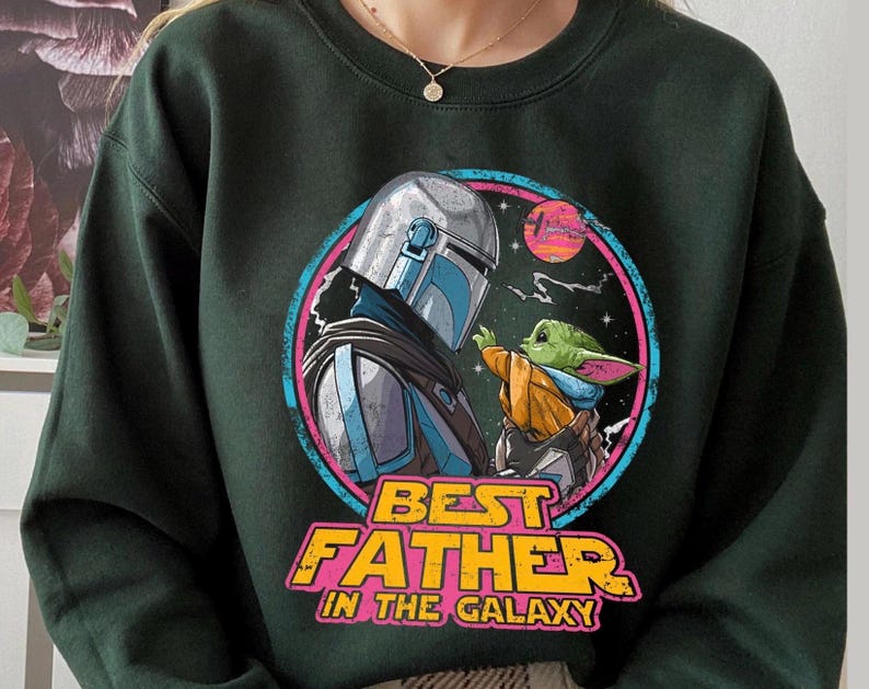 May include: Dark green sweatshirt featuring a colorful graphic of a character in silver armor and a small green creature. The graphic is framed by a pink and blue circle with the text "BEST FATHER IN THE GALAXY" in yellow and orange.