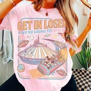 May include: Pink t-shirt with the text "GET IN LOSER WE'RE GOING TO SPACE" in orange and blue. The graphic features a space-themed amusement park ride with cartoon characters, planets, and flying saucers.