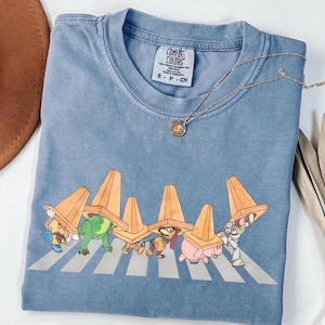 May include: A blue Comfort Colors t-shirt with a graphic of cartoon characters crossing a zebra crossing, each wearing an orange traffic cone. A gold necklace with a pendant is also visible. The shirt is folded on a white surface.