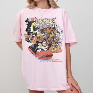 May include: A pale pink t-shirt with a vibrant graphic from the Pirates of the Caribbean attraction at Tokyo Disneyland. The design showcases Mickey Mouse, Donald Duck, and a pirate ship. The text "Pirates of the Caribbean" and "Tokyo Disneyland" are also present.