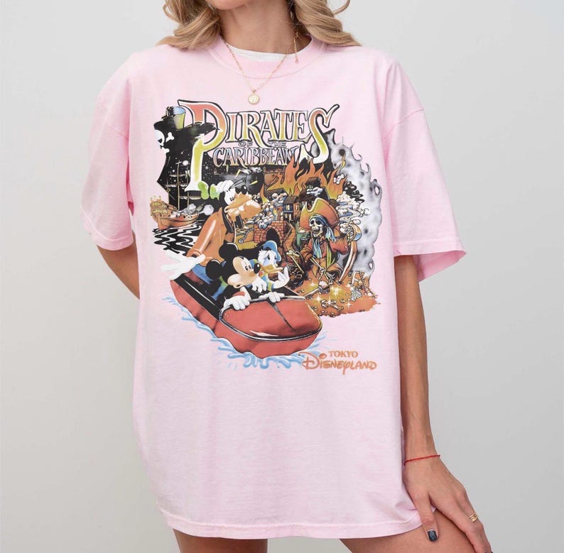 May include: A pale pink t-shirt with a vibrant graphic from the Pirates of the Caribbean attraction at Tokyo Disneyland. The design showcases Mickey Mouse, Donald Duck, and a pirate ship. The text "Pirates of the Caribbean" and "Tokyo Disneyland" are also present.