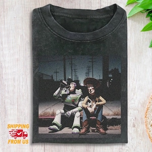 May include: Dark grey t-shirt with a distressed image of Buzz Lightyear and Woody from Toy Story. The characters are shown sitting on a kerb, with Buzz drinking from a can. The background depicts a street scene at night.