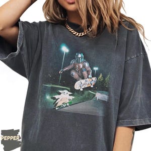 May include: Dark gray t-shirt featuring a graphic of The Mandalorian skateboarding, with Grogu on a skateboard below. The design includes a night scene with streetlights and trees. The shirt has a vintage, worn look.