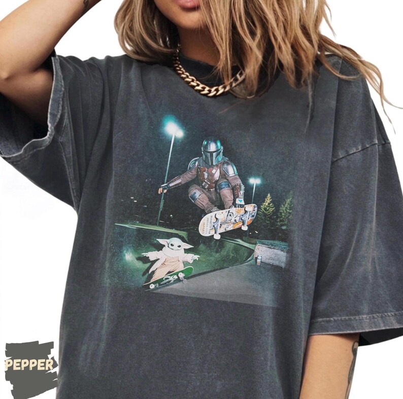 May include: Dark gray t-shirt featuring a graphic of The Mandalorian skateboarding, with Grogu on a skateboard below. The design includes a night scene with streetlights and trees. The shirt has a vintage, worn look.