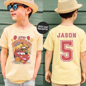 May include: A light yellow t-shirt featuring a birthday-themed graphic with the text "It's My Birthday" and a cartoon car design. The back of the shirt displays the name "Jason" and the number "5" in red.