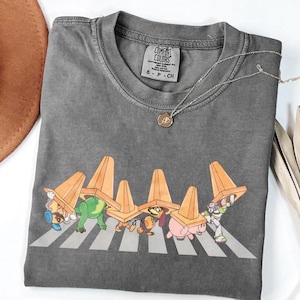 May include: Grey Comfort Colors t-shirt with a cartoon graphic of Toy Story characters crossing a zebra crossing, each wearing an orange traffic cone. A silver necklace is also visible.