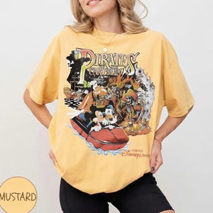 May include: Mustard yellow t-shirt with a graphic of the Pirates of the Caribbean ride at Tokyo Disneyland. The design includes Mickey Mouse, Donald Duck, and a pirate ship. The shirt has short sleeves and a crew neck.