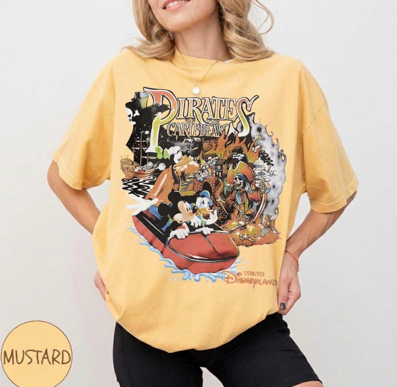May include: Mustard yellow t-shirt with a graphic of the Pirates of the Caribbean ride at Tokyo Disneyland. The design includes Mickey Mouse, Donald Duck, and a pirate ship. The shirt has short sleeves and a crew neck.