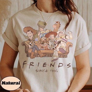 May include: Beige t-shirt featuring a graphic of Toy Story characters sitting on a couch. The word "FRIENDS" is printed above "SINCE 1995". The shirt has rolled-up sleeves and a "Natural" label.