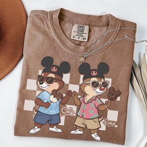 Peut inclure: T-shirt marron Comfort Colors avec une illustration de Chip et Dale portant des lunettes de soleil et tenant des glaces. Le t-shirt a un motif &agrave; carreaux et le mot "Chip" est imprim&eacute;.