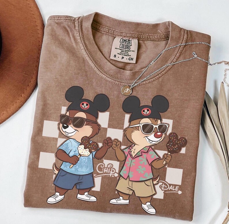 Peut inclure: T-shirt marron Comfort Colors avec une illustration de Chip et Dale portant des lunettes de soleil et tenant des glaces. Le t-shirt a un motif &agrave; carreaux et le mot "Chip" est imprim&eacute;.