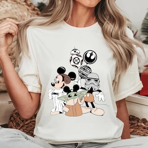 May include: Cream-colored t-shirt featuring a graphic of Mickey Mouse, Minnie Mouse, Baby Yoda, and Star Wars characters. The design includes BB-8, a Stormtrooper, and the Rebel Alliance and Galactic Empire symbols.