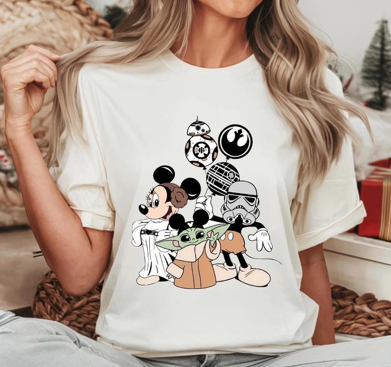 May include: Cream-colored t-shirt featuring a graphic of Mickey Mouse, Minnie Mouse, Baby Yoda, and Star Wars characters. The design includes BB-8, a Stormtrooper, and the Rebel Alliance and Galactic Empire symbols.