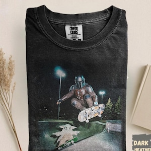 May include: Dark heather gray t-shirt featuring a graphic of The Mandalorian and Grogu skateboarding at night. The shirt has the Comfort Colors label at the neckline. The graphic is in shades of gray, brown, and green.