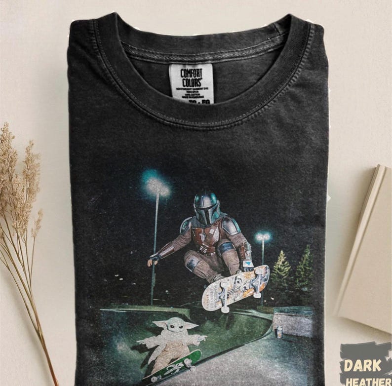 May include: Dark heather gray t-shirt featuring a graphic of The Mandalorian and Grogu skateboarding at night. The shirt has the Comfort Colors label at the neckline. The graphic is in shades of gray, brown, and green.
