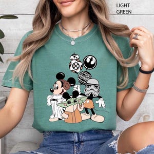 May include: Light green t-shirt featuring a graphic of Mickey Mouse, Baby Yoda, and Star Wars characters. The design includes a Stormtrooper, BB-8, and other iconic elements from the Star Wars franchise.