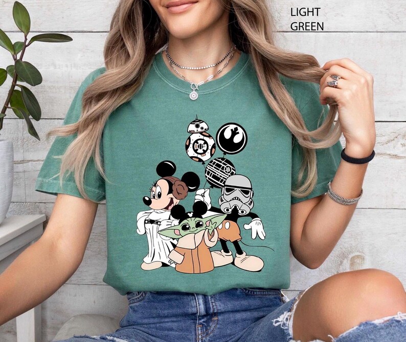 May include: Light green t-shirt featuring a graphic of Mickey Mouse, Baby Yoda, and Star Wars characters. The design includes a Stormtrooper, BB-8, and other iconic elements from the Star Wars franchise.