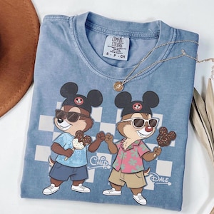 Peut inclure: Un t-shirt bleu Comfort Colors avec un dessin anim&eacute; de Tic et Tac portant des lunettes de soleil et tenant des glaces. Le t-shirt a un fond &agrave; carreaux et les mots "Chip" et "Dale" sont imprim&eacute;s sur le t-shirt. Un collier en or est drap&eacute; sur le t-shirt.