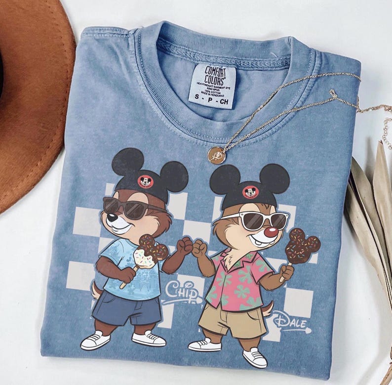 Peut inclure: Un t-shirt bleu Comfort Colors avec un dessin anim&eacute; de Tic et Tac portant des lunettes de soleil et tenant des glaces. Le t-shirt a un fond &agrave; carreaux et les mots "Chip" et "Dale" sont imprim&eacute;s sur le t-shirt. Un collier en or est drap&eacute; sur le t-shirt.