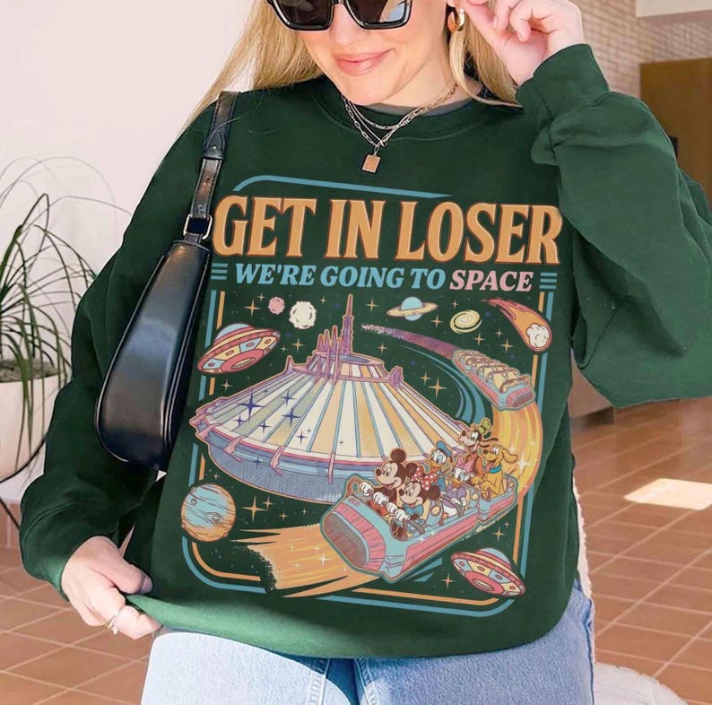 May include: Dark green sweatshirt with the text "GET IN LOSER WE'RE GOING TO SPACE" and a colorful graphic of a space-themed amusement park ride with cartoon characters. The sweatshirt has long sleeves.