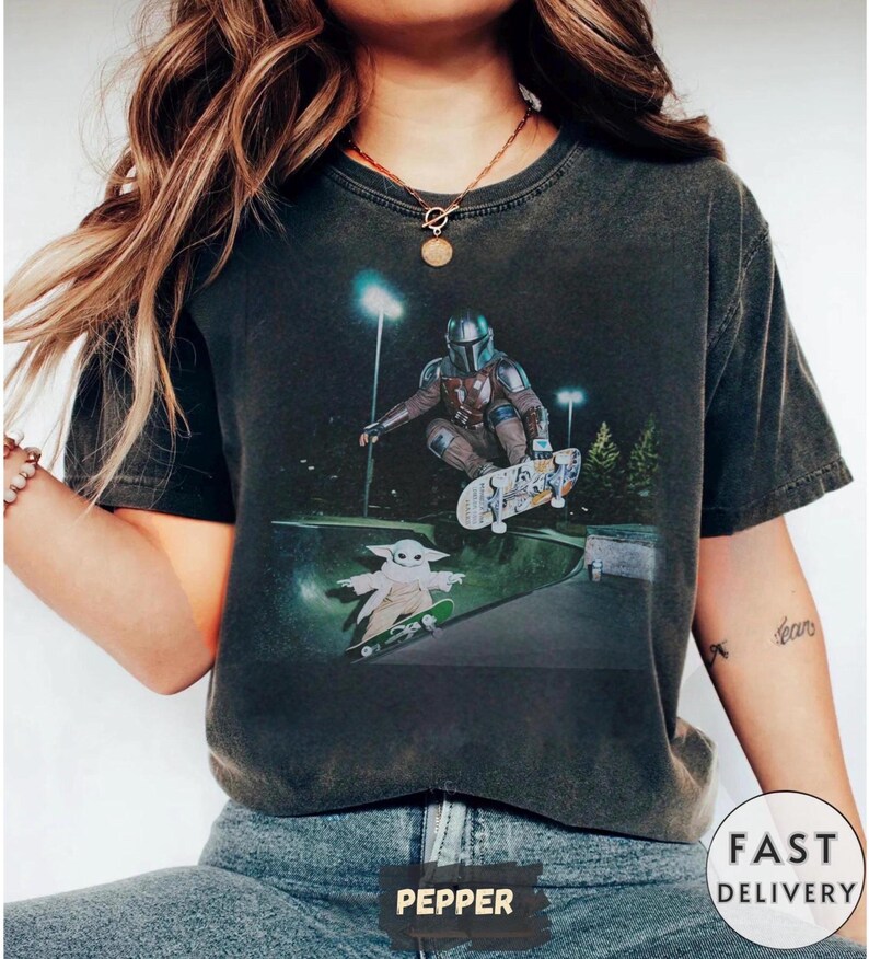 May include: Dark gray t-shirt featuring a graphic of a character in armor skateboarding above a ramp, with a smaller character on a skateboard below. The design is set against a night scene with lights and trees.