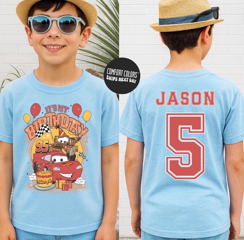 May include: A light blue t-shirt featuring a birthday design with the text "It's My Birthday" and a cartoon car graphic. The back of the shirt displays the name "Jason" and the number "5" in red. The shirt is made of Comfort Colors material.